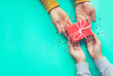 Two pairs of hands are captured in the act of giving and receiving a small, red polka-dotted gift box tied with white string. The turquoise background is sprinkled with colorful confetti, adding to the festive and celebratory mood of the scene.