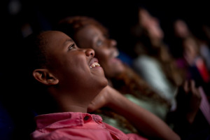 Boy in movie theater looking up at screen and smiling, with other patrons in background