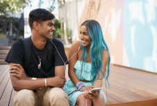 A couple looks at each other smiling while listening to music through earbuds outdoors