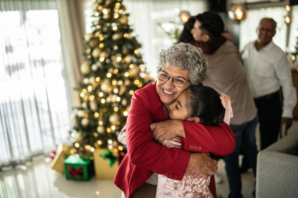 Grandmother hugging granddaughter, with Christmas tree and family in the background