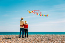 The back of two older people arm in arm on the beach, with a colorful kite in the sky