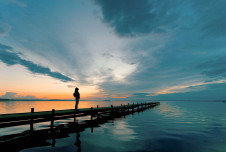 Person standing alone on a dock by the water at dusk, with clouds