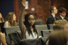 In a conference setting, a young woman with braided hair, wearing a brown sweater, raises her hand to participate. She sits among other attendees who appear focused on the session, with some of them wearing formal attire. The background shows a professional environment with a mixture of attention and engagement from the participants.