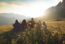 Four people with a dog, from behind, sitting on a grassy hill with mountains and sunlight in the background