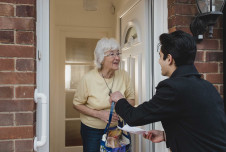 A young man delivers groceries to an elderly woman at her front door; they smile and engage in a pleasant conversation.