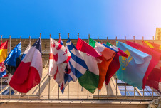 Flags of various countries are displayed on flagpoles in front of a building under a clear blue sky