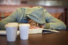 Exhausted young woman with her head on a book on a desk, and multiple cups of coffee in front of her