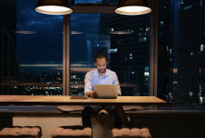 A man working late at night in a modern office with a laptop, surrounded by city lights visible through large windows