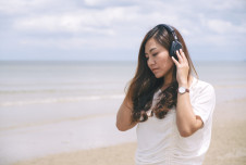 A woman listens to music with headphones while standing on a beach, appearing contemplative