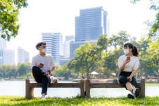 Two people wearing masks sit on a bench, engaging in conversation with a cityscape in the background