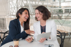 Two women in business attire are laughing and joking around at work, one holding a tablet and the other a smartphone