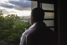 A man stands in profile, wearing a face mask, looking out the window at a sunset over a lush landscape, in a moment of quiet reflection.