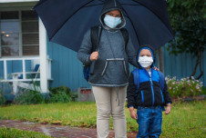 A mother and her young son, both wearing masks and rain gear, stand under an umbrella outside their house.