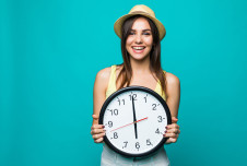 A woman in a hat holding a large clock, smiling against a teal background