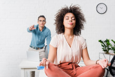 A woman is meditating with her eyes closed in an office, while a coworker in the background points and looks frustrated