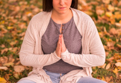 A woman is meditating outdoors with a sad expression, sitting among fallen leaves with her hands in a prayer position