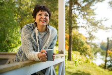 A woman stands on a porch, holding a coffee cup and smiling, with a backdrop of trees and greenery
