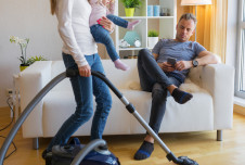 A woman is vacuuming the floor while holding a small child in her arms. In the background, a man is sitting on a white couch, looking at his phone.