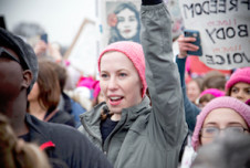 A woman in a pink hat raises her fist during the Women's March on Washington, surrounded by other participants and protest signs