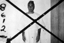Black and white photo showing a man dressed in a white robe, standing against a wall with the number "8612" beside him. A large "X" is drawn over the image.