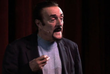 Philip Zimbardo speaking on stage, wearing a dark blazer and a light gray shirt