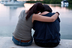 Two people sitting side by side on a dock on a lake, one embracing the other, from behind