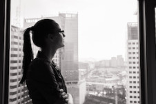 A woman with glasses stands by a window, gazing thoughtfully at a cityscape