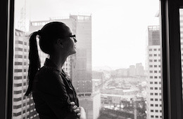 A woman with glasses stands by a window, gazing thoughtfully at a cityscape