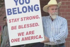 A man wearing a cowboy hat and striped shirt holds a large sign that reads: "YOU BELONG. STAY STRONG. BE BLESSED. WE ARE ONE AMERICA."